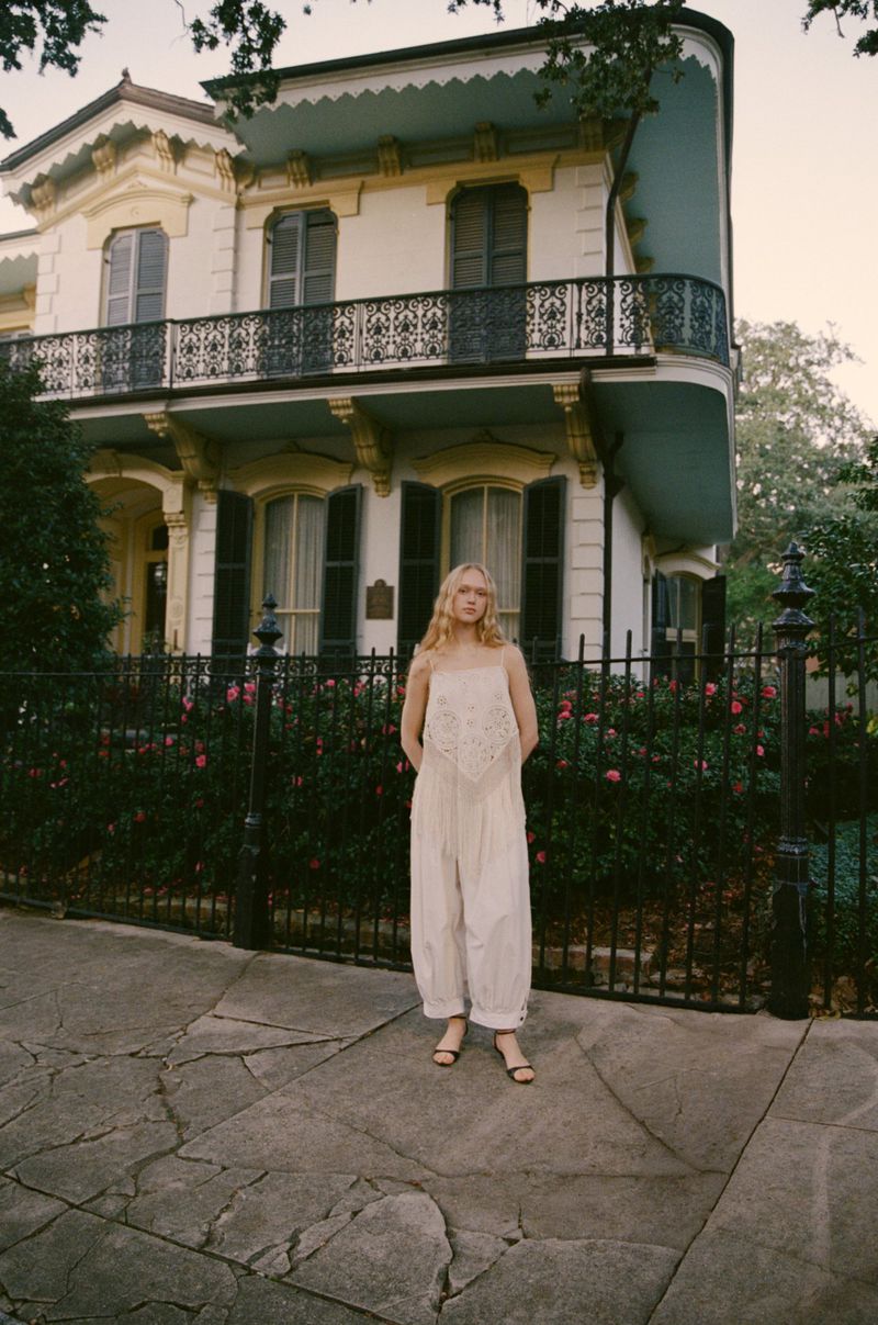 A woman in a white jumpsuit stands in front of a large, classic house with a black iron fence and blooming pink roses, creating a soft, elegant scene.