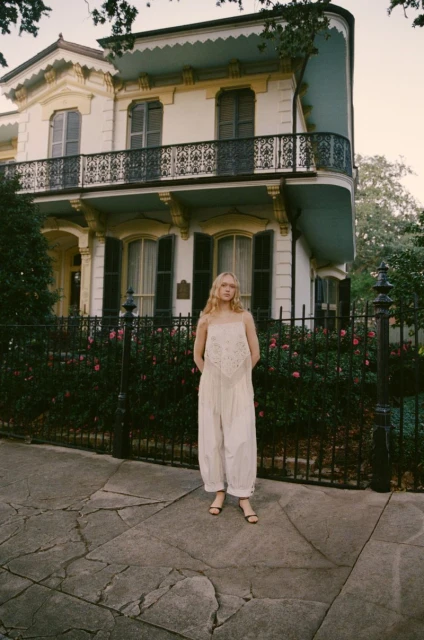 A woman in a white jumpsuit stands in front of a large, classic house with a black iron fence and blooming pink roses, creating a soft, elegant scene.