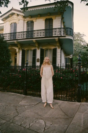 A woman in a white jumpsuit stands in front of a large, classic house with a black iron fence and blooming pink roses, creating a soft, elegant scene.