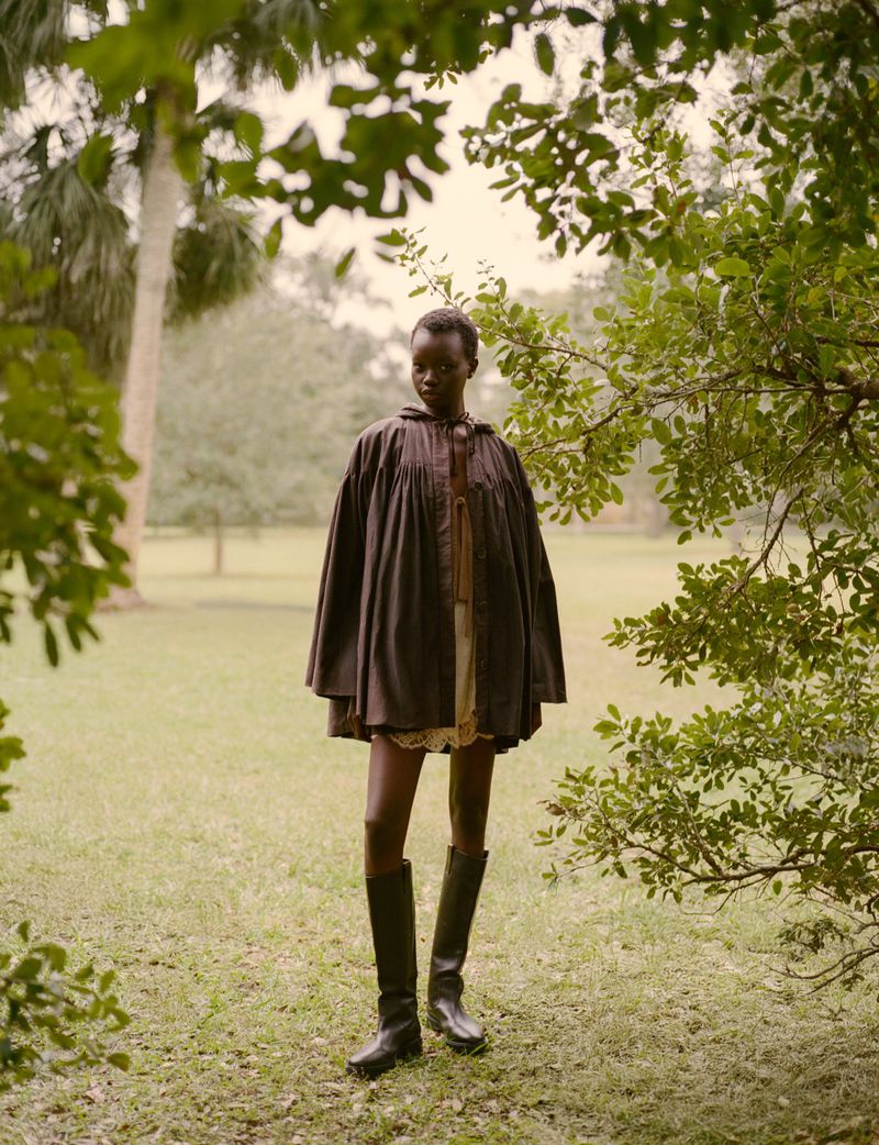 Model wearing a dark smock coat and tall leather boots standing outdoors in a grassy park framed by tree foliage.