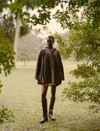 Model wearing a dark smock coat and tall leather boots standing outdoors in a grassy park framed by tree foliage.