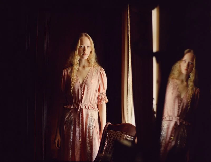 Young woman with long blonde hair posing in a pink dress with a reflection from a mirror inside a dimly lit room with dark wood.