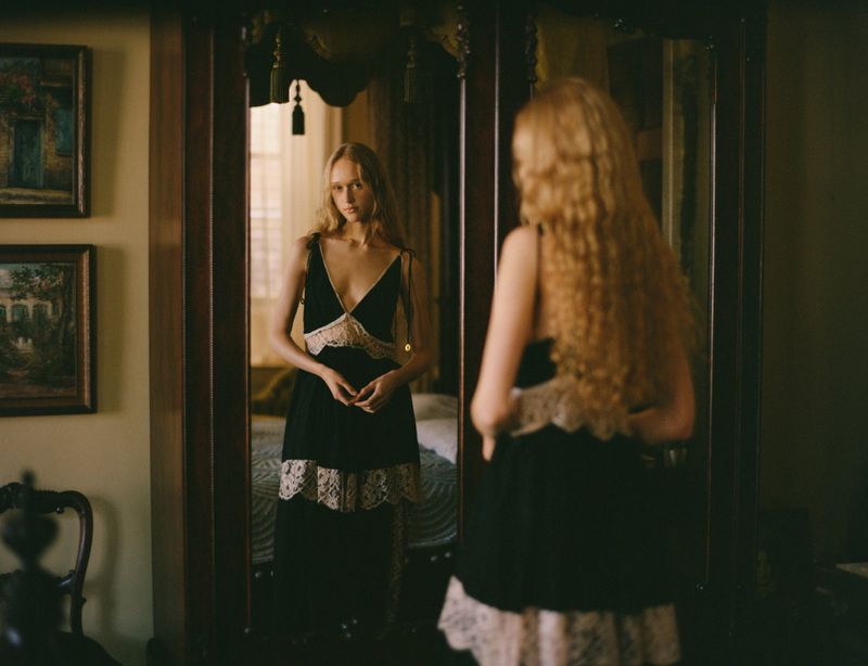 Woman in a black lace-trimmed dress stands in a bedroom, reflected in a wooden wardrobe mirror; soft indoor lighting.