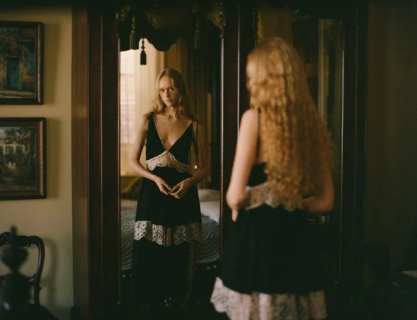 Woman in a black lace-trimmed dress stands in a bedroom, reflected in a wooden wardrobe mirror; soft indoor lighting.