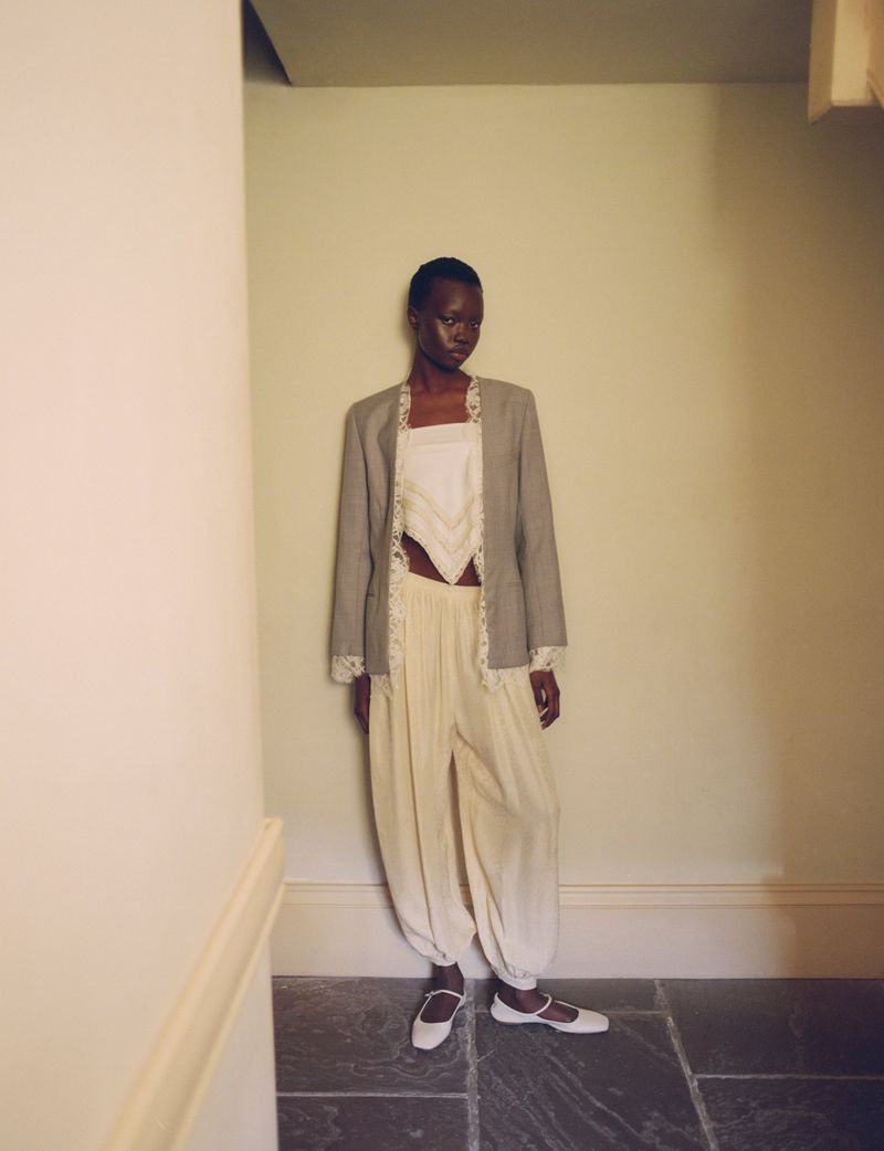 Fashion shot of a slender Black woman posing indoors, wearing a grey blazer, white top, cream pants, and flats against a neutral wall.