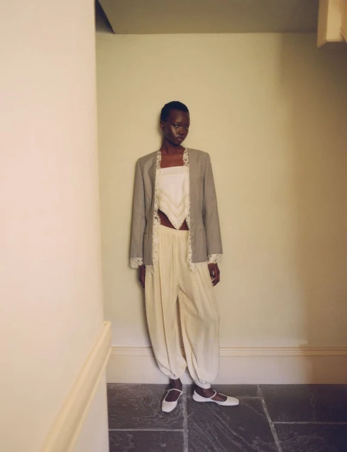 Fashion shot of a slender Black woman posing indoors, wearing a grey blazer, white top, cream pants, and flats against a neutral wall.