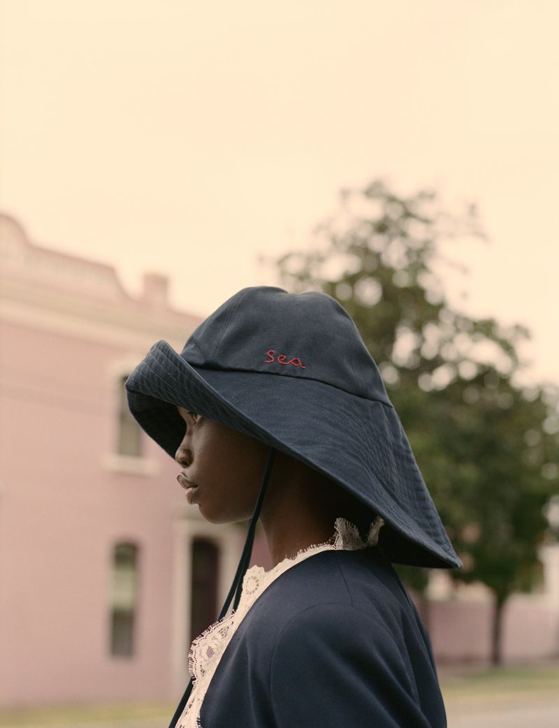 Elegant portrait of a Black woman in a wide-brimmed hat and navy blazer against a backdrop of pink buildings and green trees.