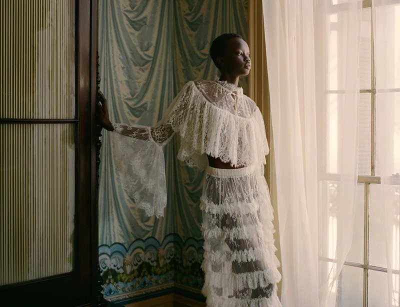Elegant African-American woman poses in white lace gown by a window, bringing together fashion, beauty, and artistic photography.