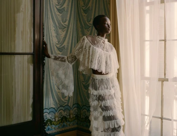 Elegant African-American woman poses in white lace gown by a window, bringing together fashion, beauty, and artistic photography.