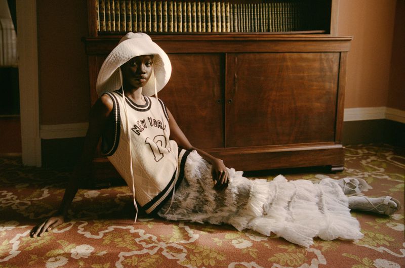 A woman lies on a floral carpet wearing a wide-brimmed hat, a knit basketball jersey, and a white ruffled skirt, with a wooden cabinet behind.