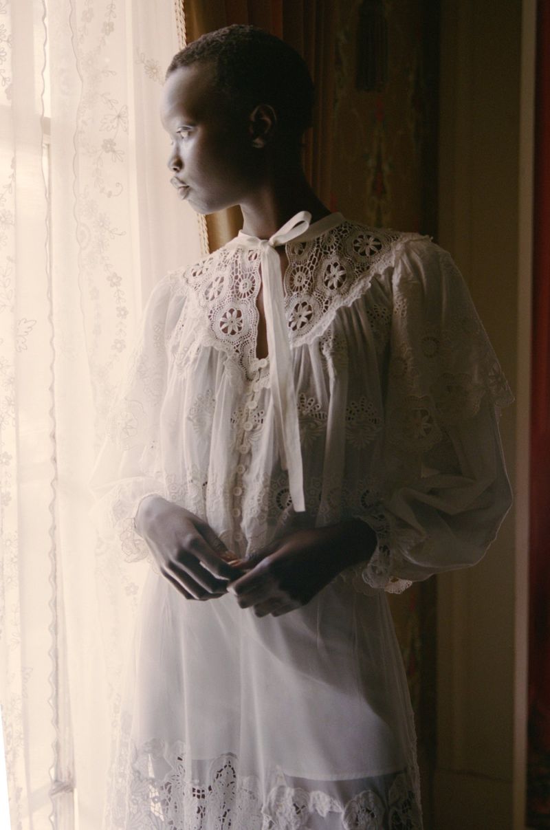 Elegant Black woman in white lace nightgown stands by a window with natural light, highlighting her soft features and timeless beauty.