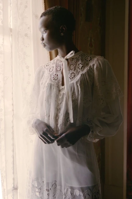 Elegant Black woman in white lace nightgown stands by a window with natural light, highlighting her soft features and timeless beauty.