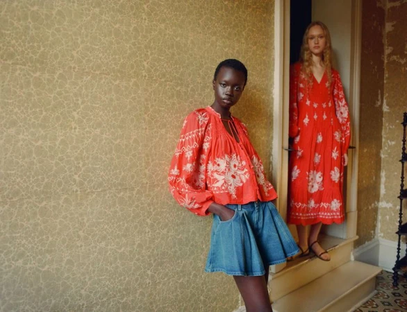 Two models wearing red embroidered folk garments and denim shorts posing by an ornate textured wall and doorway.