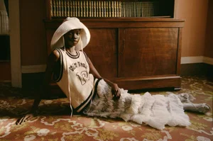 Model wearing a wide-brimmed white hat, knit tank top, and ruffled skirt reclining on an ornate patterned carpet in a room