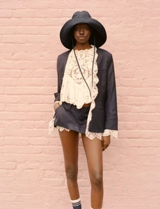 Model wearing a wide-brimmed black hat, dark blazer, lace top, and shorts standing against a pink brick wall.