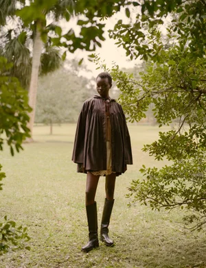 Model wearing a dark smock coat and tall leather boots standing outdoors in a grassy park framed by tree foliage.