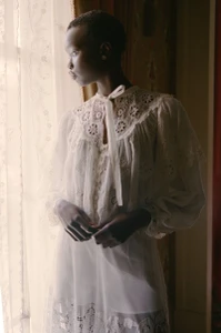 Model standing by a window wearing a white lace blouse and dress, looking out into natural light.