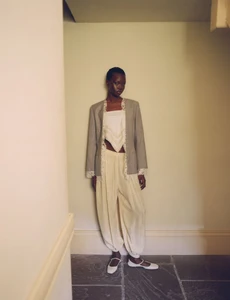 Model Aker Ajak standing in a domestic interior wearing a grey blazer, cream top, and light trousers.