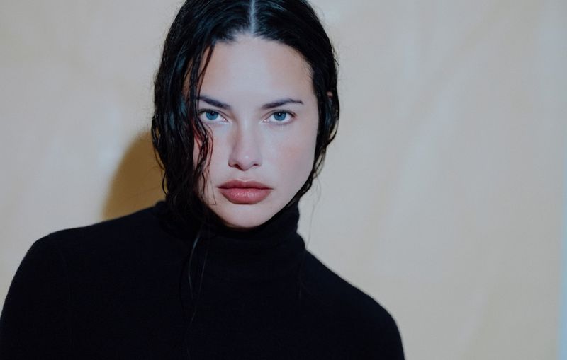 Close-up portrait of a woman with slicked-back dark hair and blue eyes, wearing a black turtleneck against a muted yellow background.