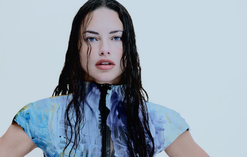 Close-up portrait of a woman with wet dark hair wearing a blue, white, and purple paint-patterned short-sleeved top against a white background.
