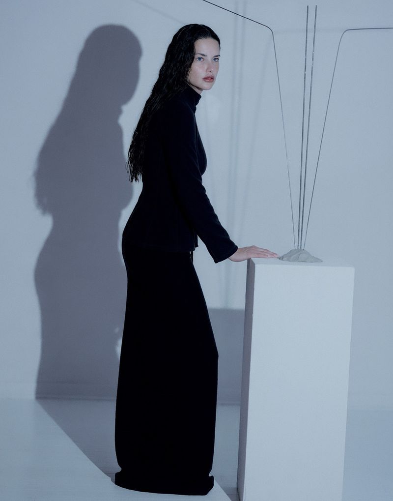 A woman stands in a dark long-sleeved dress and maxi skirt next to a geometric object in a brightly lit indoor photo studio.