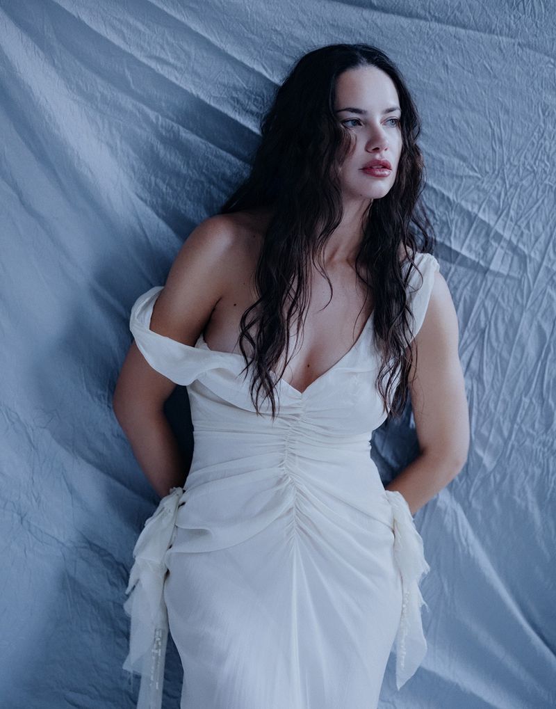 Glamorous woman in a white off-the-shoulder dress poses against a draped blue backdrop, creating a dreamy, elegant aesthetic.