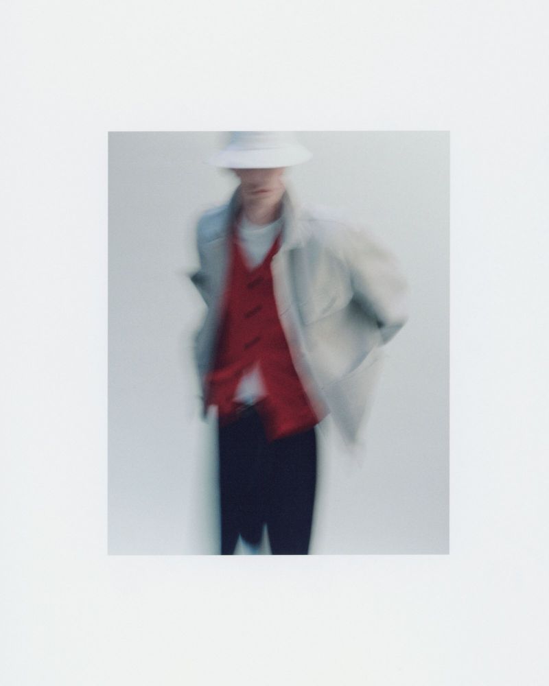 Blurred fashion photo of a man in a bucket hat, red vest, and coat, posing in a photo studio with soft lighting against a white backdrop.