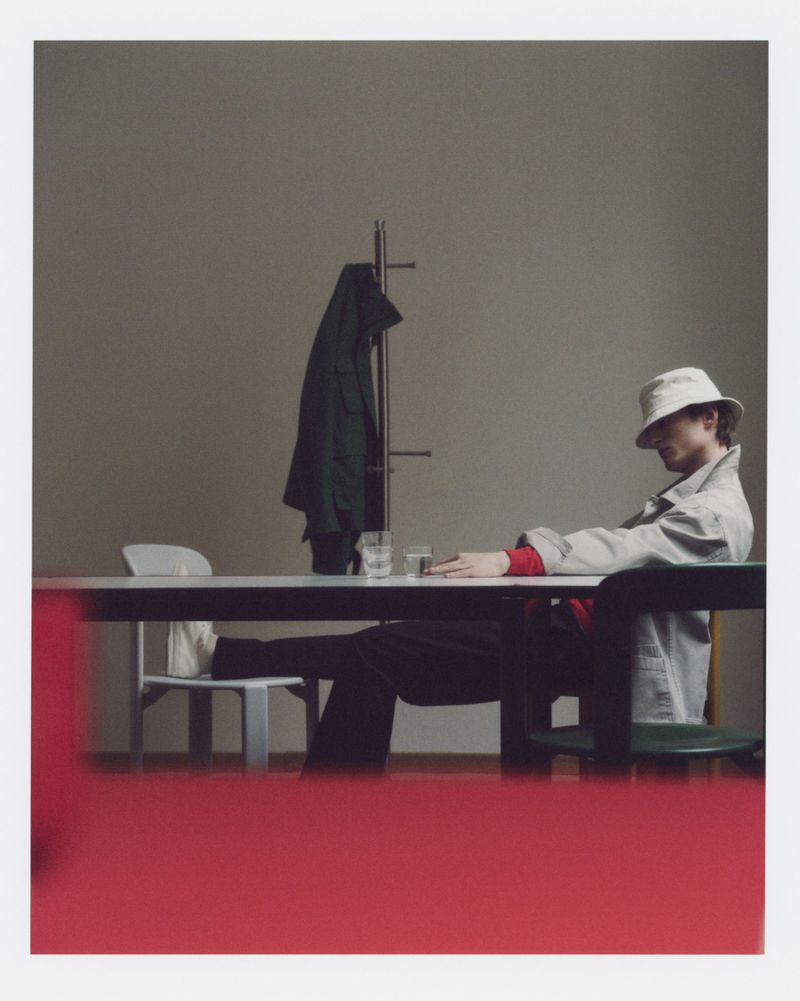 A person sitting at a table wearing a bucket hat and jacket in a simple room with a coat rack and minimal decor.