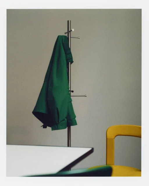 Green jacket hangs on a metal coat rack near a yellow chair and white table in a brightly lit indoor setting with neutral wall paint.