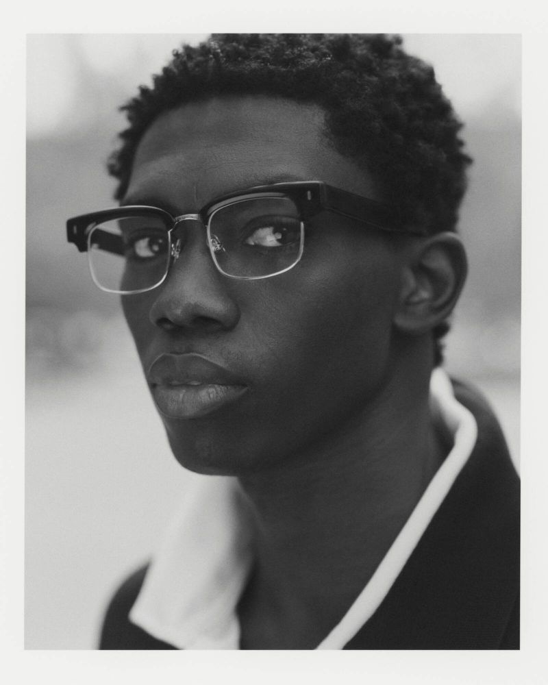 Monochromatic portrait of a young adult Black male in eyeglasses and a jacket with a white collar, looking directly at the camera.