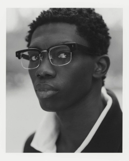 Monochromatic portrait of a young adult Black male in eyeglasses and a jacket with a white collar, looking directly at the camera.