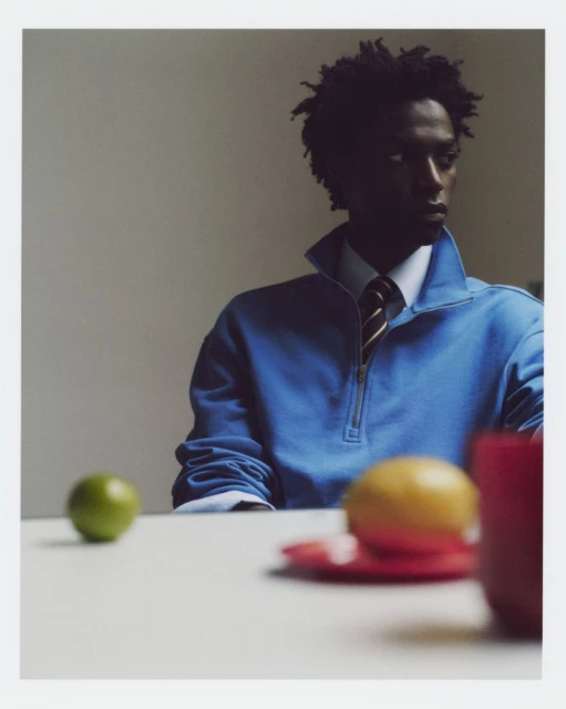 Young man in blue zip-up sweater sits at a table with fruit, creating a minimalist, indoor scene with soft, natural lighting.