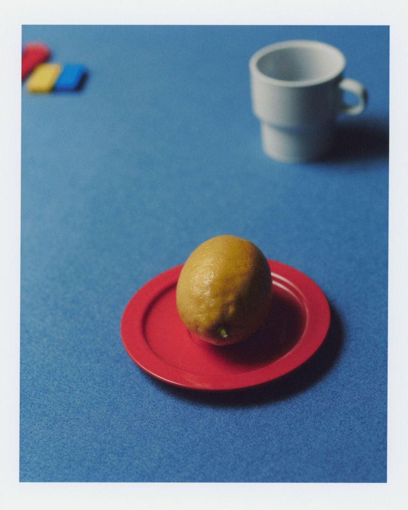 A lemon sits on a red plate with a white cup and colorful blocks on a blue surface in a brightly lit indoor setting.