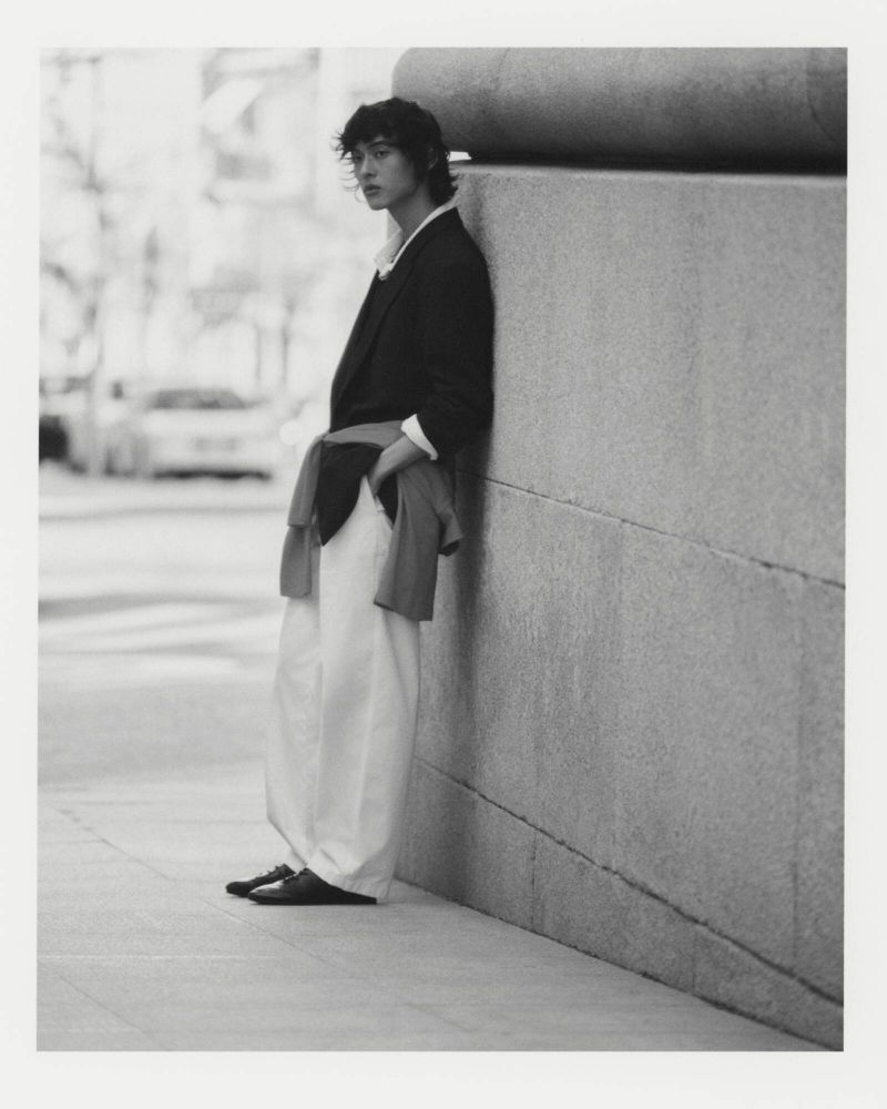Young adult man in a blazer and white pants stands against a stone wall in an urban setting, captured in a black and white portrait.