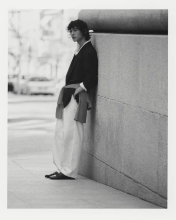 Young adult man in a blazer and white pants stands against a stone wall in an urban setting, captured in a black and white portrait.
