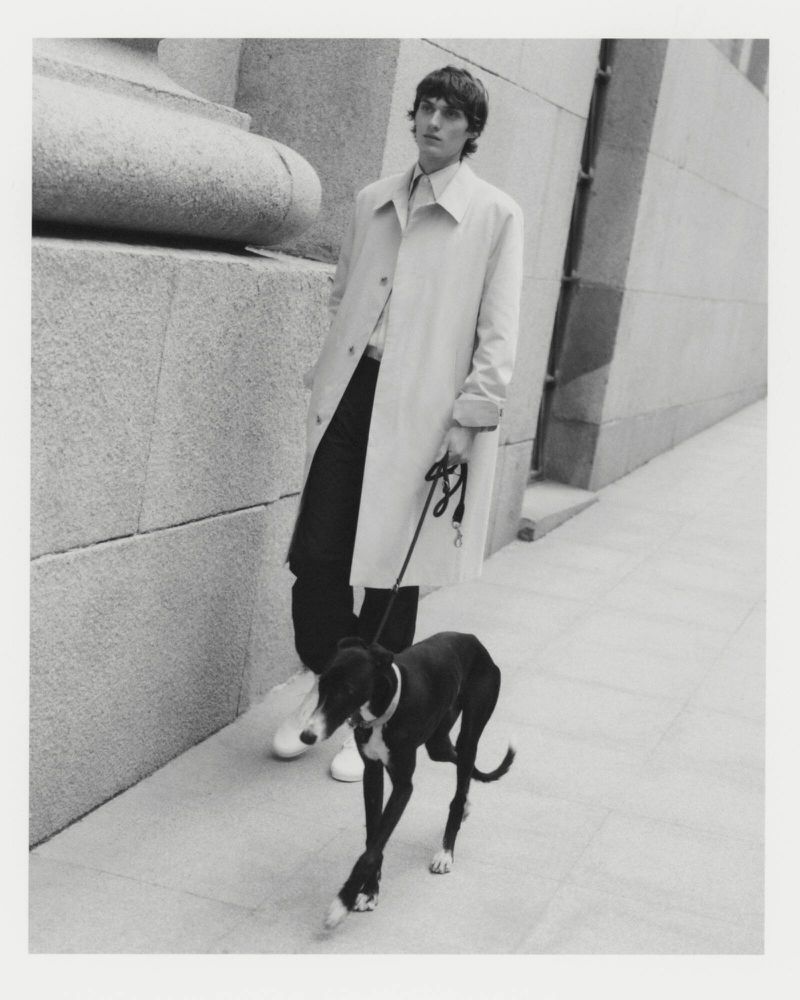 Monochrome fashion shot of a slender man in a trench coat walking a black dog on a leash along a city sidewalk near a stone building.