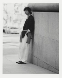 Model leaning against a stone wall on a city street wearing a dark blazer, white trousers, and a sweater tied around his waist