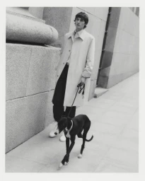 Model wearing a light coat and dark trousers walking a dog along a stone sidewalk next to a classical building exterior