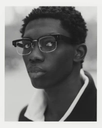 Portrait of a Black man wearing thick-rimmed glasses and a collared jacket against a neutral backdrop