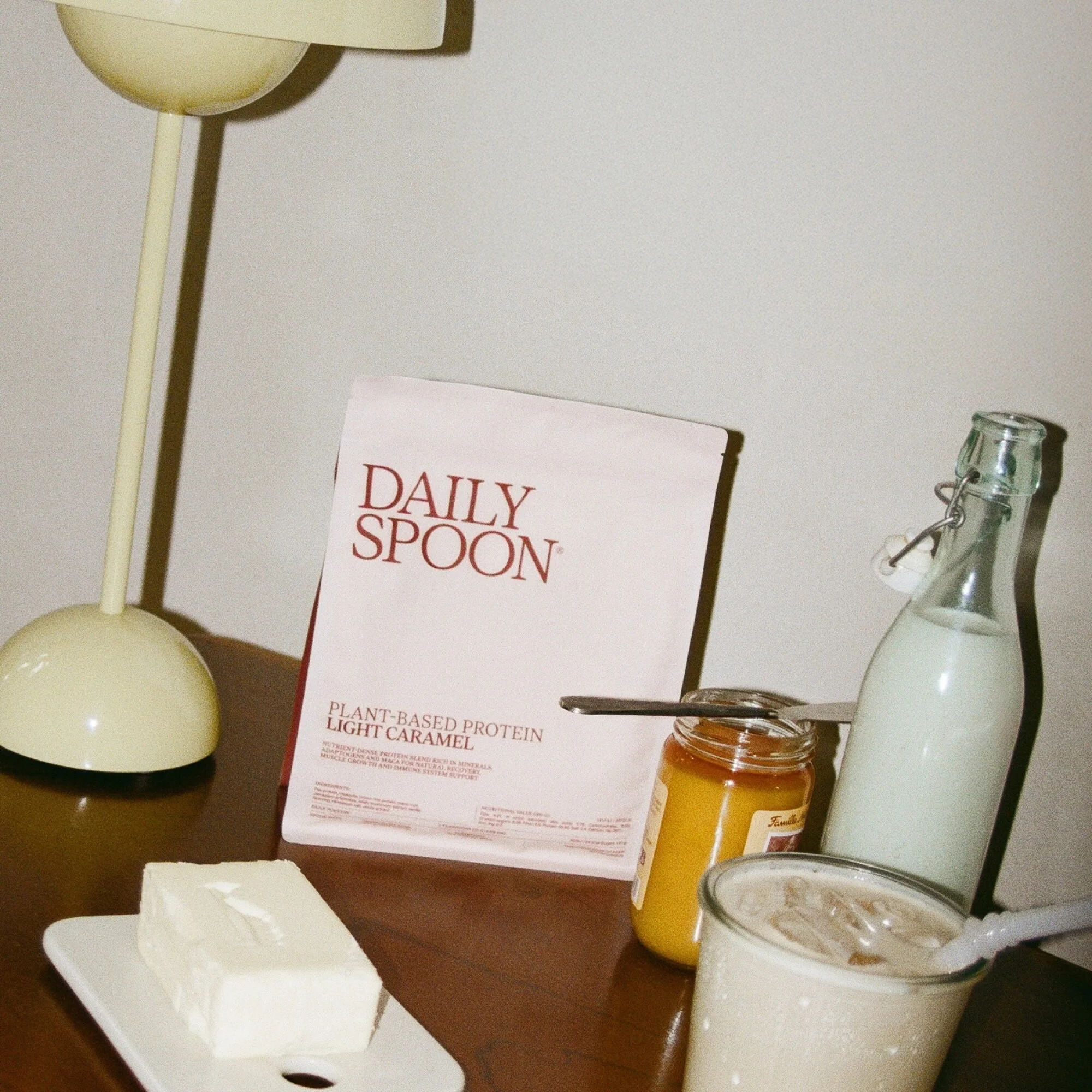Still life of Daily Spoon plant-based protein, caramel jar, coffee beverage, milk bottle, and white chocolate on wood table indoors.