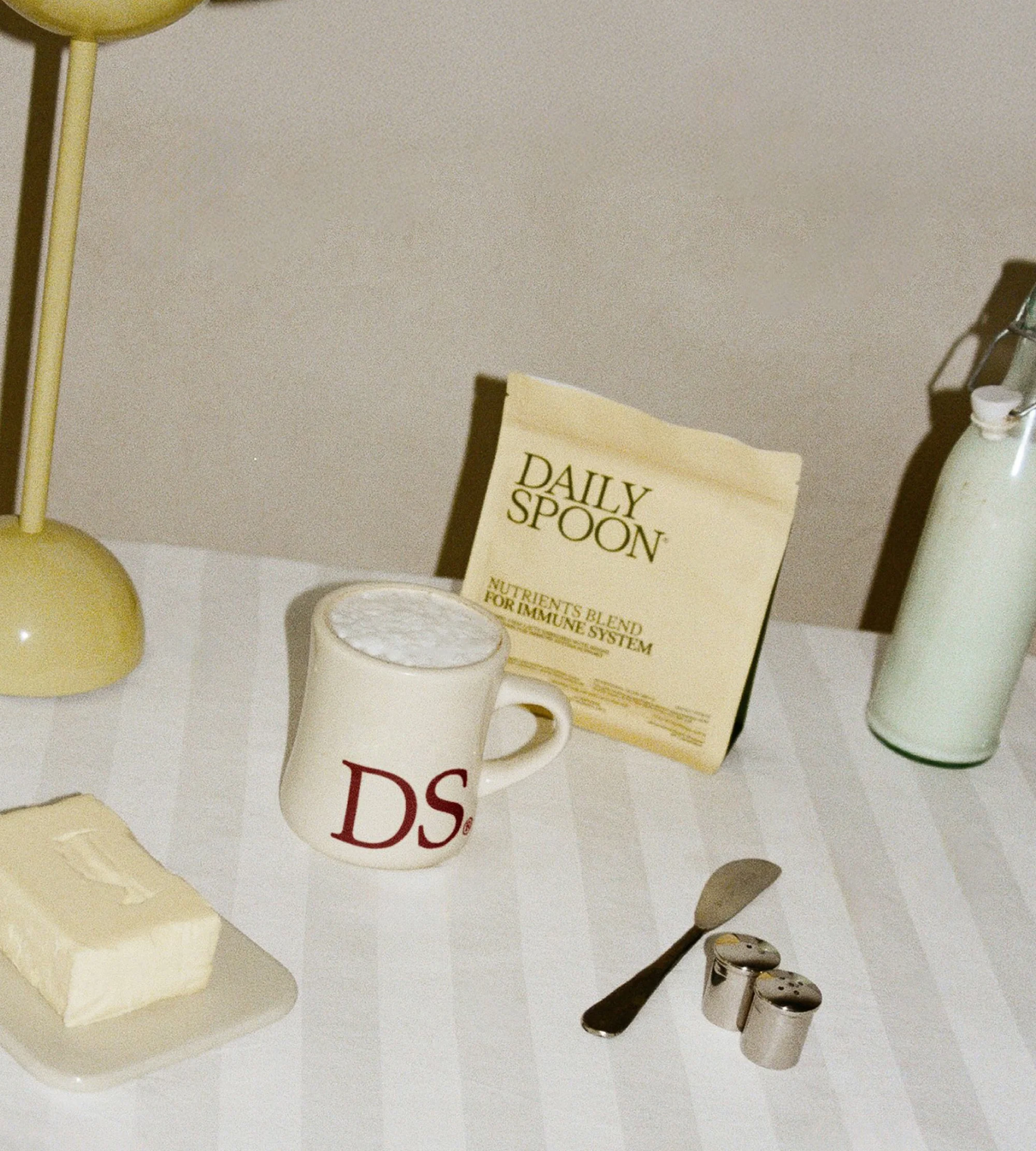 Product shot with a Daily Spoon nutrients packet, mug with frothy liquid, bottle, butter, salt and pepper shakers, and lamp on a striped cloth.