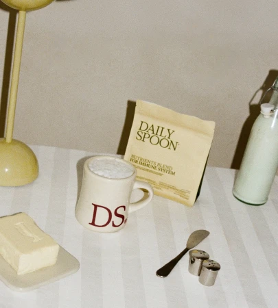 Product shot with a Daily Spoon nutrients packet, mug with frothy liquid, bottle, butter, salt and pepper shakers, and lamp on a striped cloth.