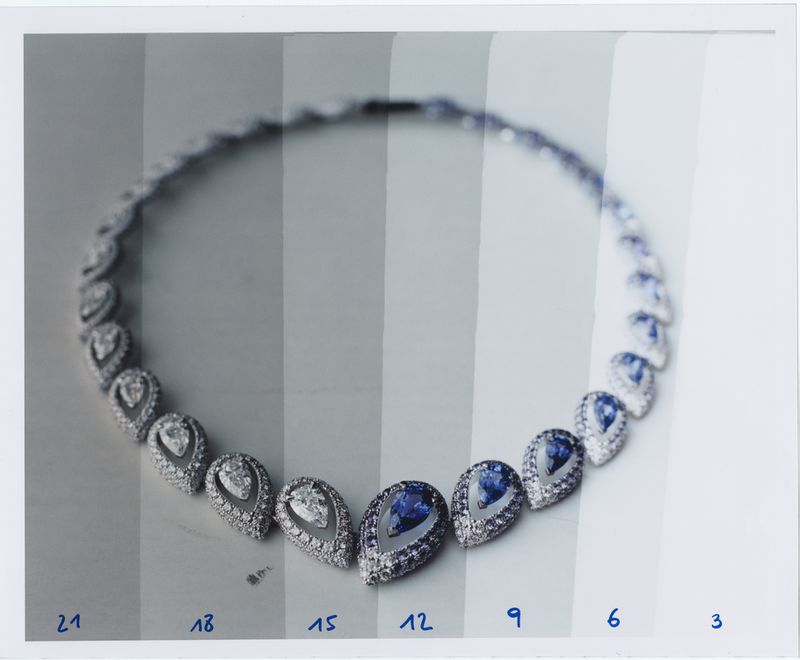 A luxury necklace featuring a repeating pattern of diamond and blue gem teardrop shapes is displayed on a graded gray backdrop.