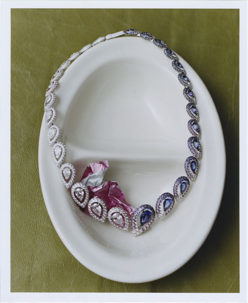 Elegant necklace with teardrop-shaped sapphires and diamonds displayed in a white dish with pink wrapping on an olive surface.