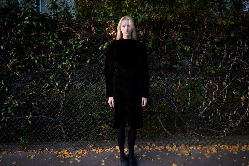 Young woman in a black coat stands in front of a leafy fence on a city street with fallen leaves, captured in a full shot.
