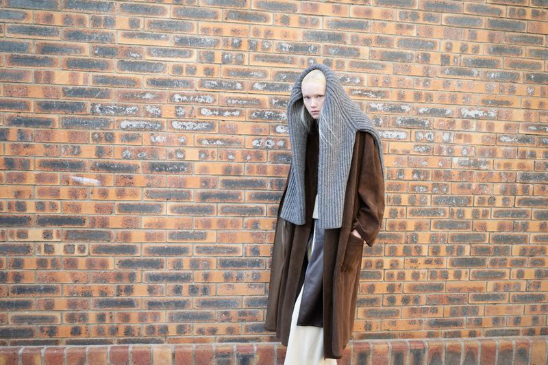 Fashion photo of a light-skinned model wearing a brown coat and gray hooded scarf standing in front of an orange brick wall outdoors.