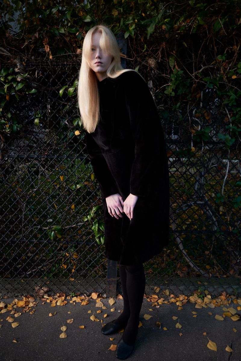 A long-haired blonde woman wearing a long black dress and black tights is posing in front of a metal fence in an outdoor setting.