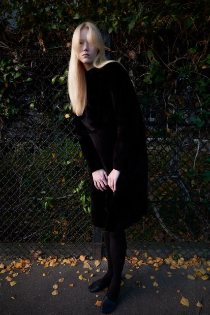 A long-haired blonde woman wearing a long black dress and black tights is posing in front of a metal fence in an outdoor setting.