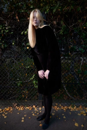A long-haired blonde woman wearing a long black dress and black tights is posing in front of a metal fence in an outdoor setting.