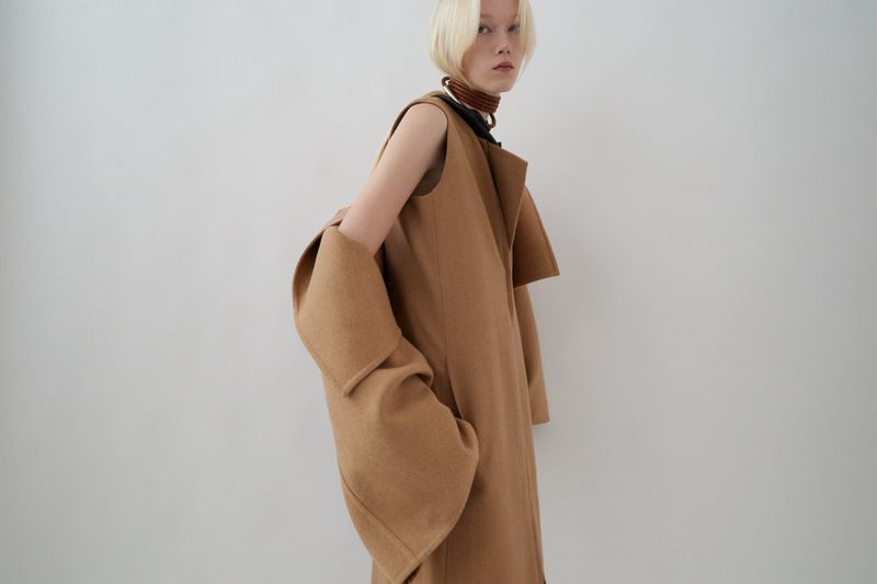 Fashion shot of a blonde woman in a tan, sleeveless coat, posing in front of a white background in a photo studio with soft lighting.